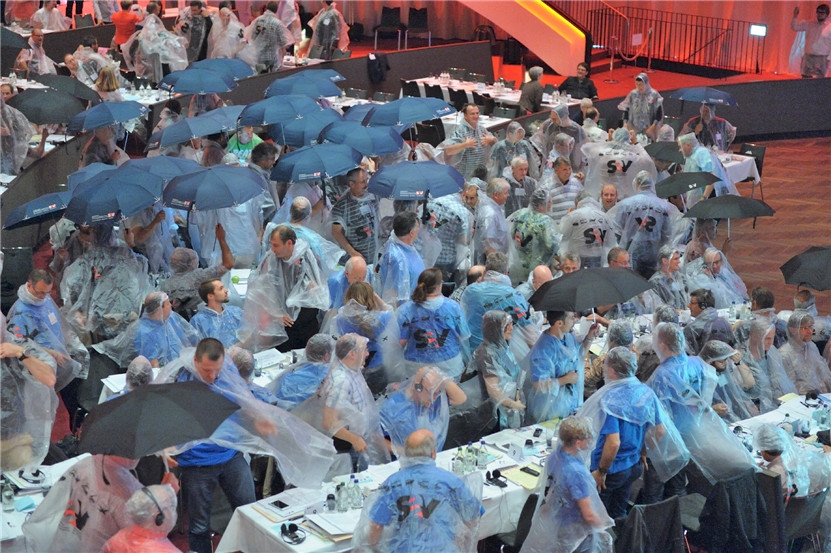 Die Delegierten ziehen Pelerinen an und spannen Schirme auf, während der BAV-Direktor auf die Tribüne steigt – aus Protest, weil er das Personal im Regen stehen lässt.